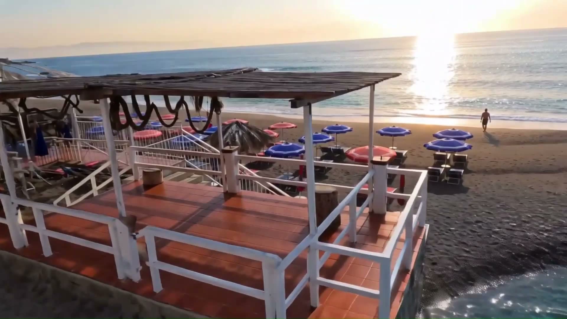 La terrazza in legno del lido al tramonto, con gli ombrelloni blu e rossi sulla sabbia scura e il mare Ionio davanti